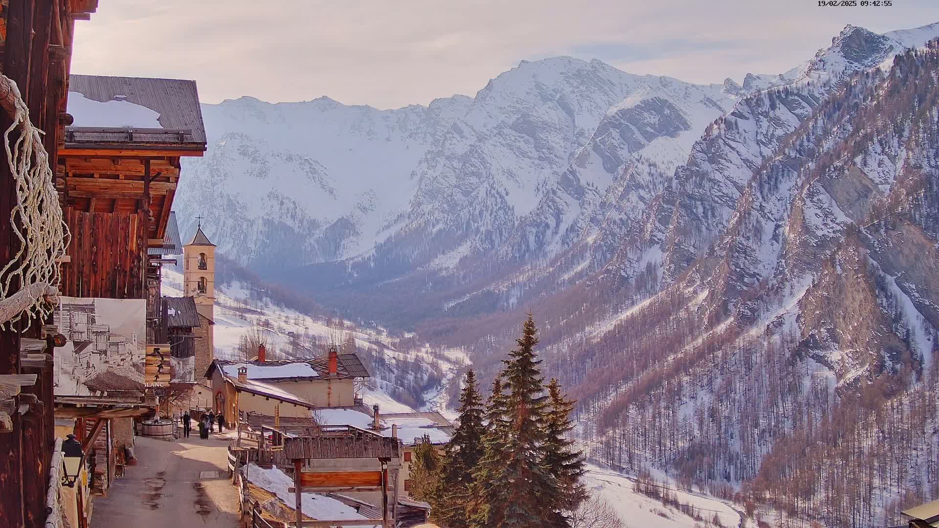 A snow-covered mountain valley is viewed from a village of rustic wooden buildings, under a partly cloudy sky.