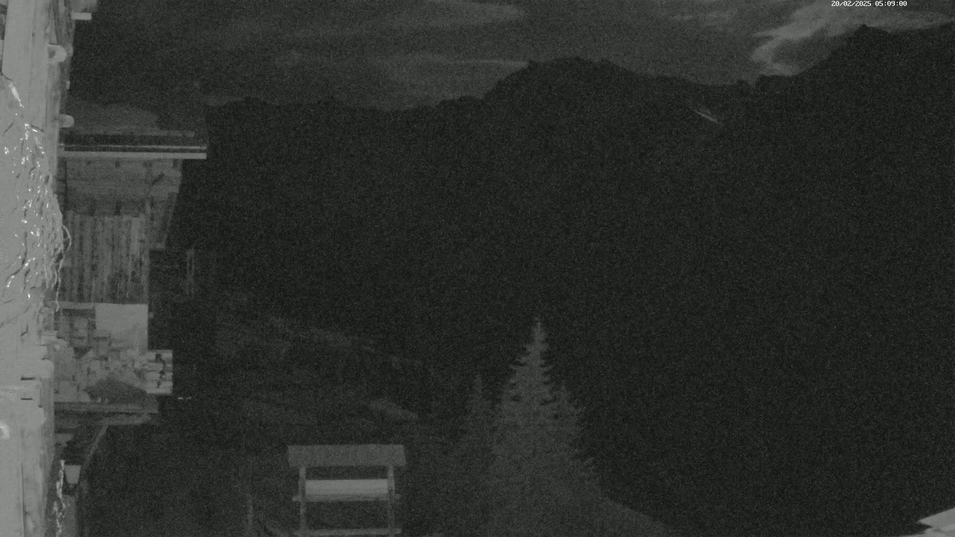 A nighttime, low-light view shows a section of a wooden building with dark mountains in the background under a cloudy sky.
