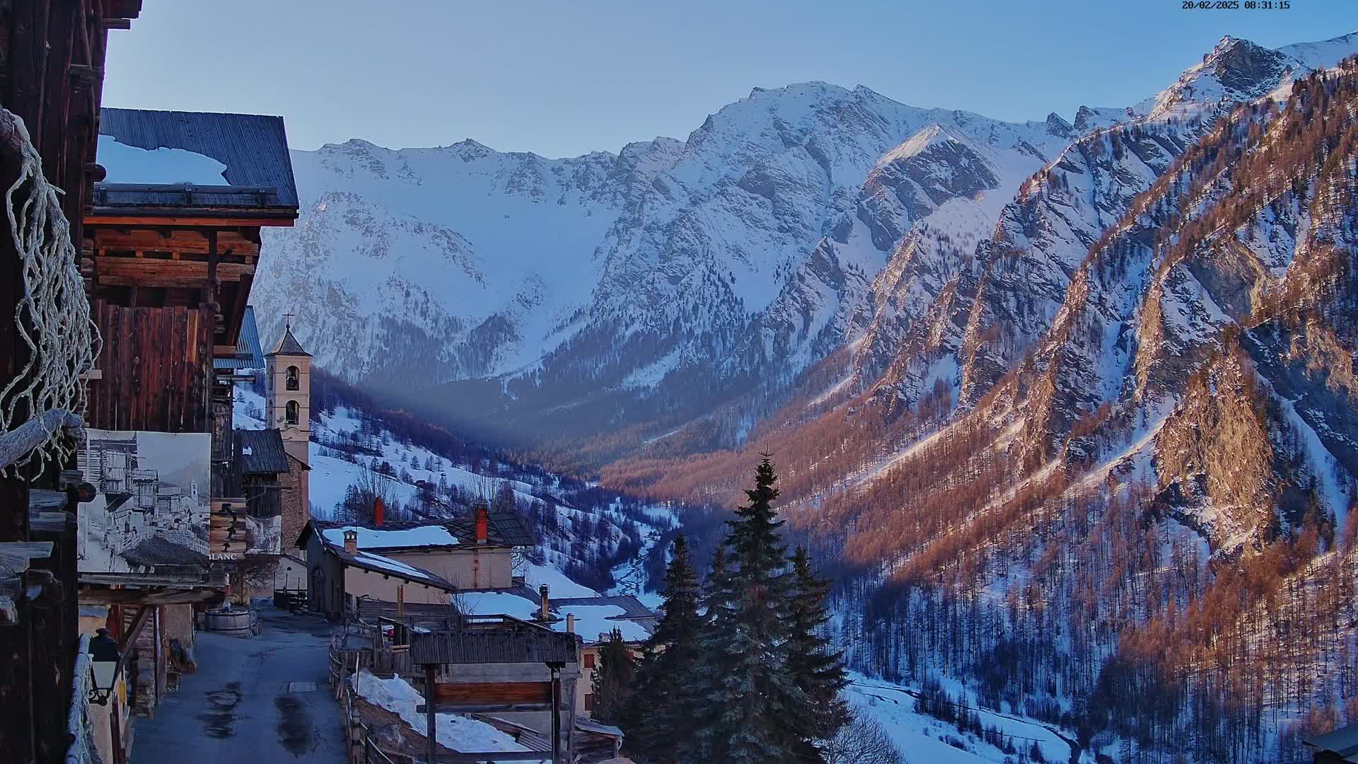 A snow-covered mountain village nestles in a valley surrounded by towering, snow-capped mountains under a clear, bright sky.