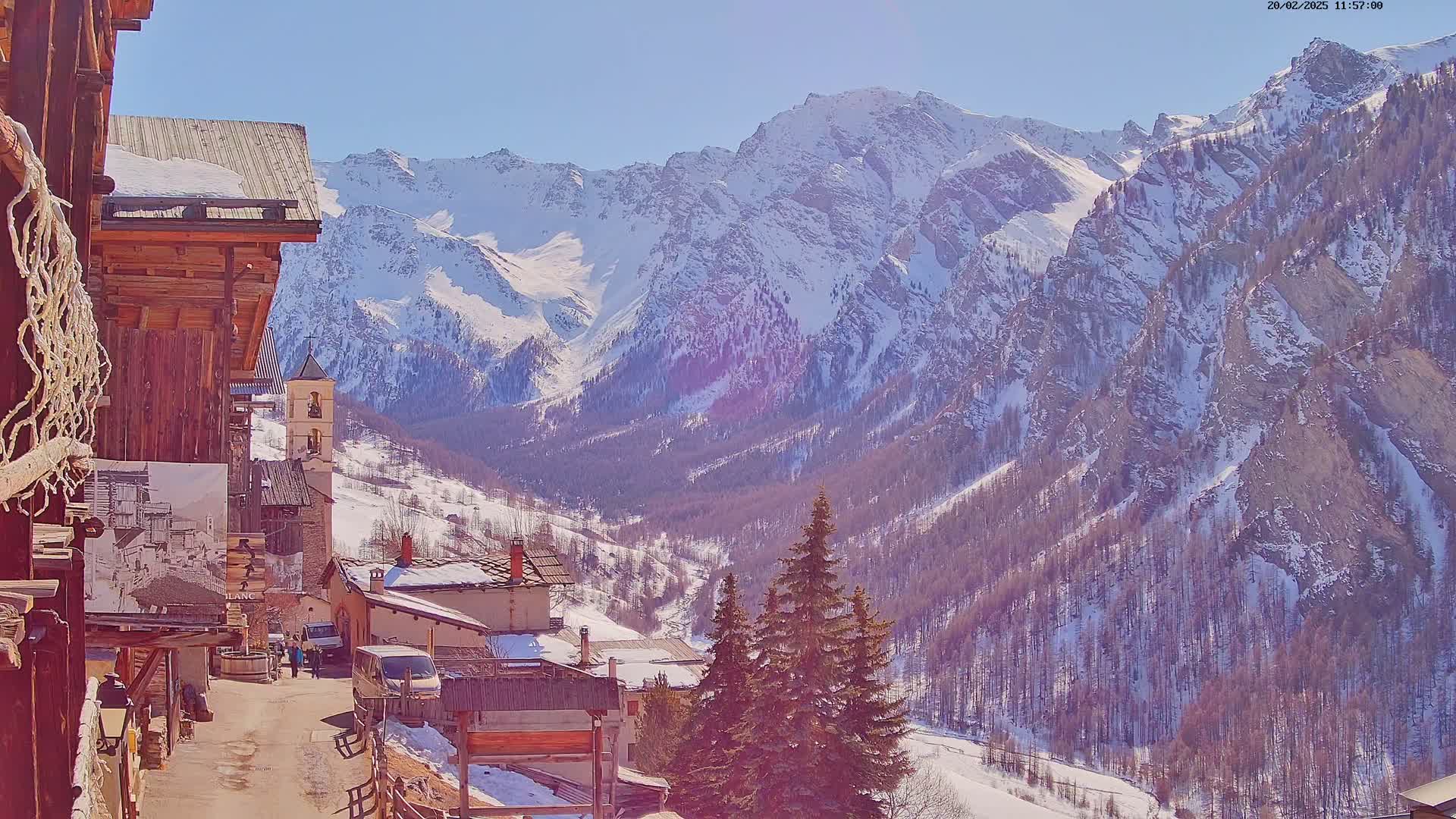 A sunny day reveals a snow-covered mountain range overlooking a quaint village nestled in a valley, with several evergreen trees in the foreground.
