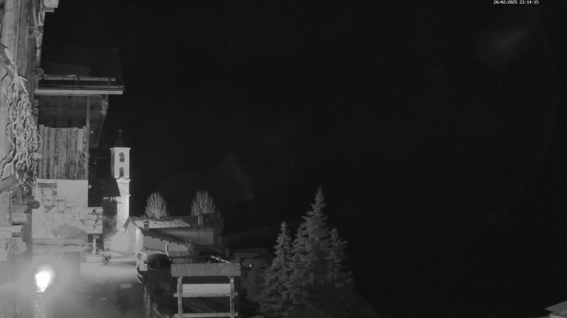 A nighttime, monochrome view of a village scene featuring a bell tower, buildings, and evergreens under a dark sky.
