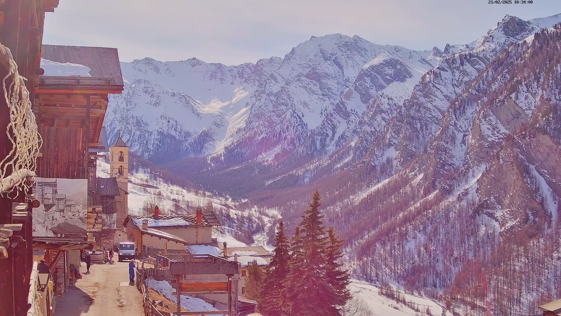 A snow-covered mountain village nestles in a valley beneath a bright, sunny sky and snow-capped mountains.