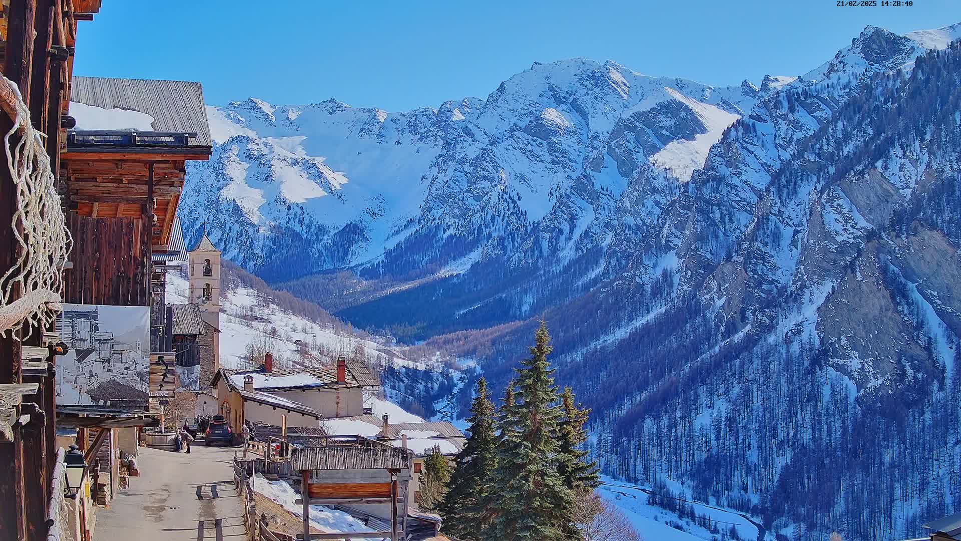 A snow-covered mountain village with a church steeple is nestled in a valley surrounded by snow-capped mountains under a clear blue sky.
