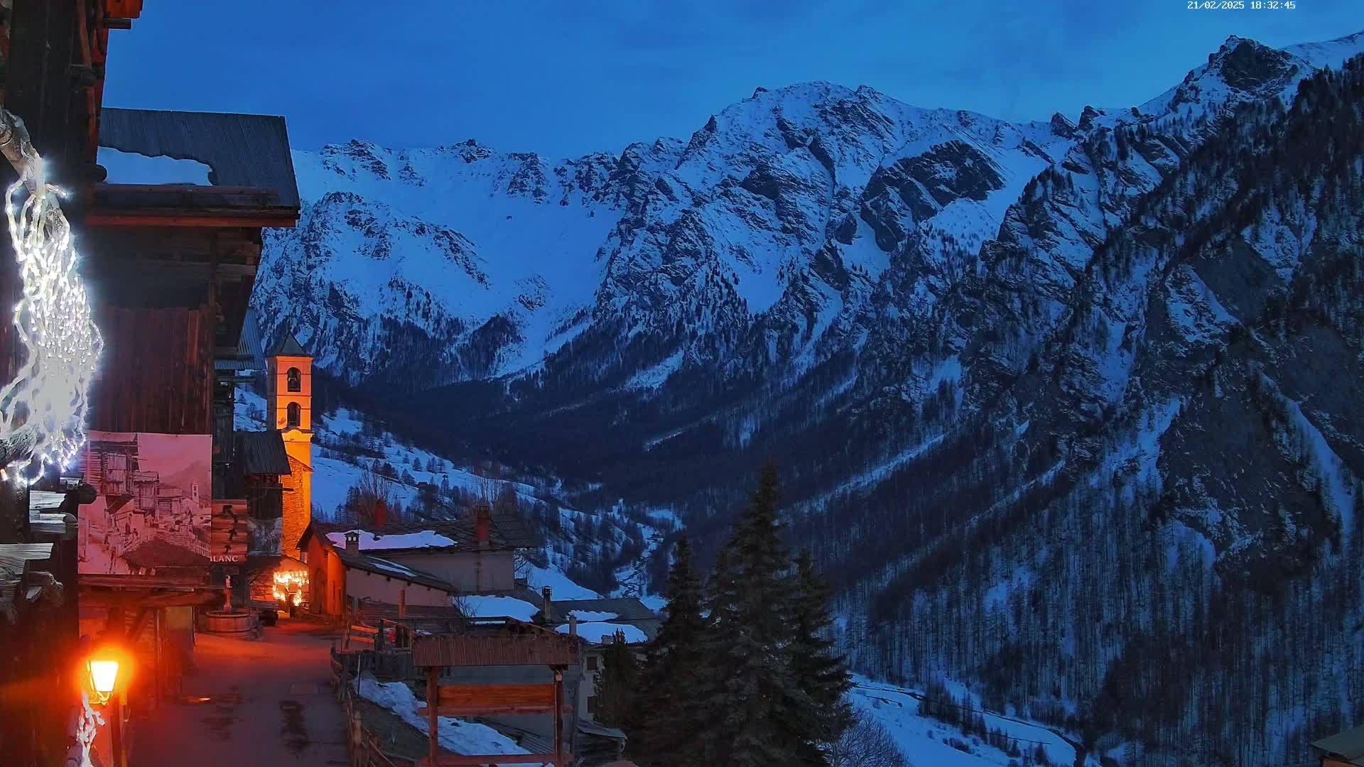 A snow-covered mountain range is visible at twilight behind a small village with illuminated buildings and strings of lights.