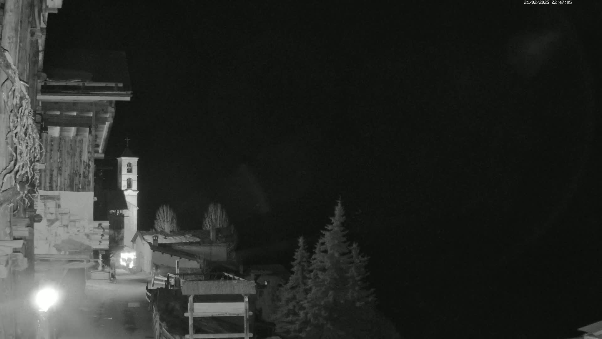 A nighttime, monochrome view of a snow-covered village street, featuring a tall bell tower and several buildings, under a dark, clear sky.