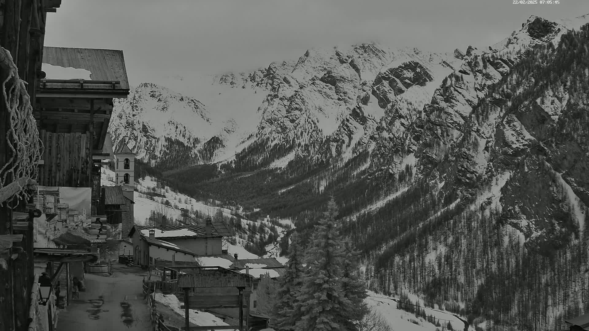 A snow-covered mountain range overlooks a small village of dark wooden buildings, under an overcast sky.