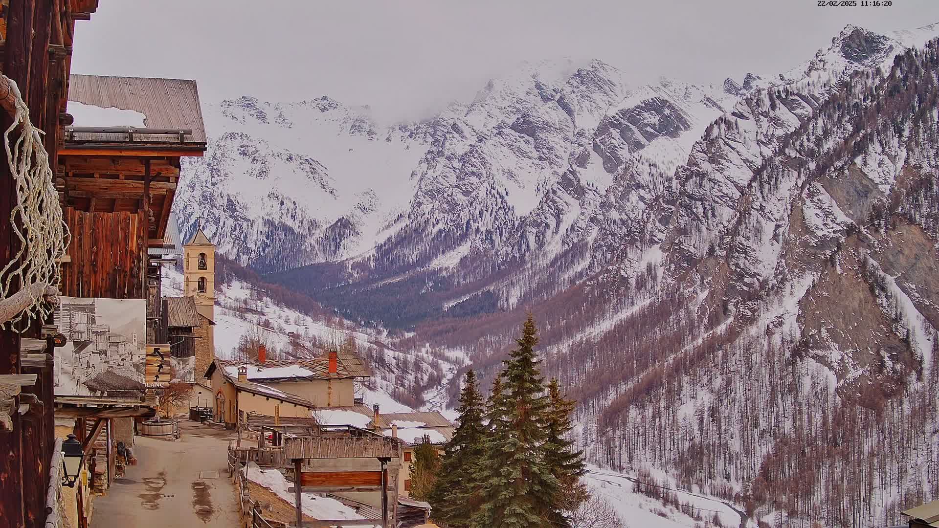A snow-covered mountain valley is viewed from a village of old wooden buildings on a cloudy day.