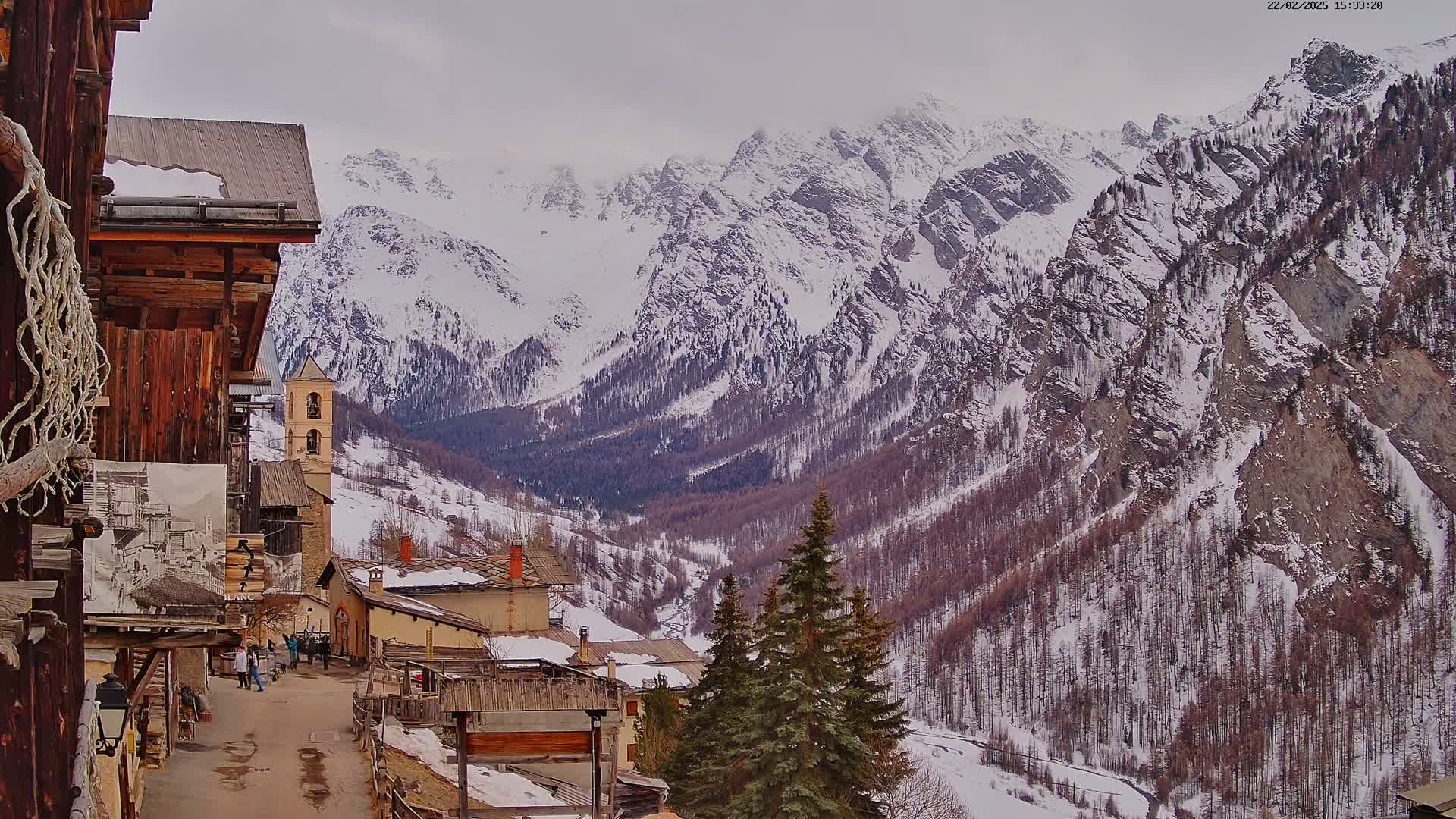 A snow-covered mountain valley is viewed from a village of rustic buildings, with overcast skies.