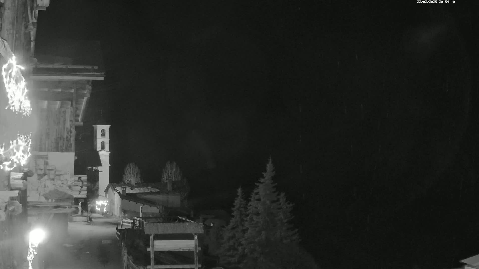 A nighttime, rain-swept view of a small village with a bell tower, partially obscured by darkness and low light.