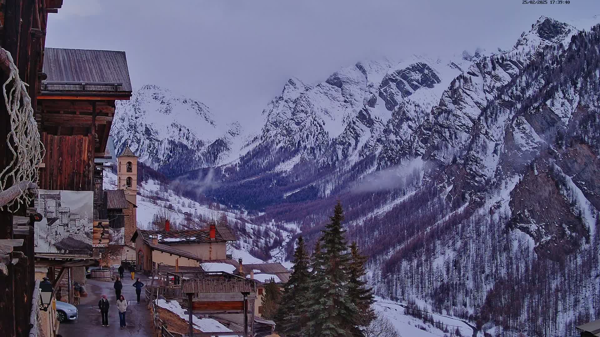 A snow-covered mountain valley is viewed from a village of old wooden buildings, under an overcast sky.