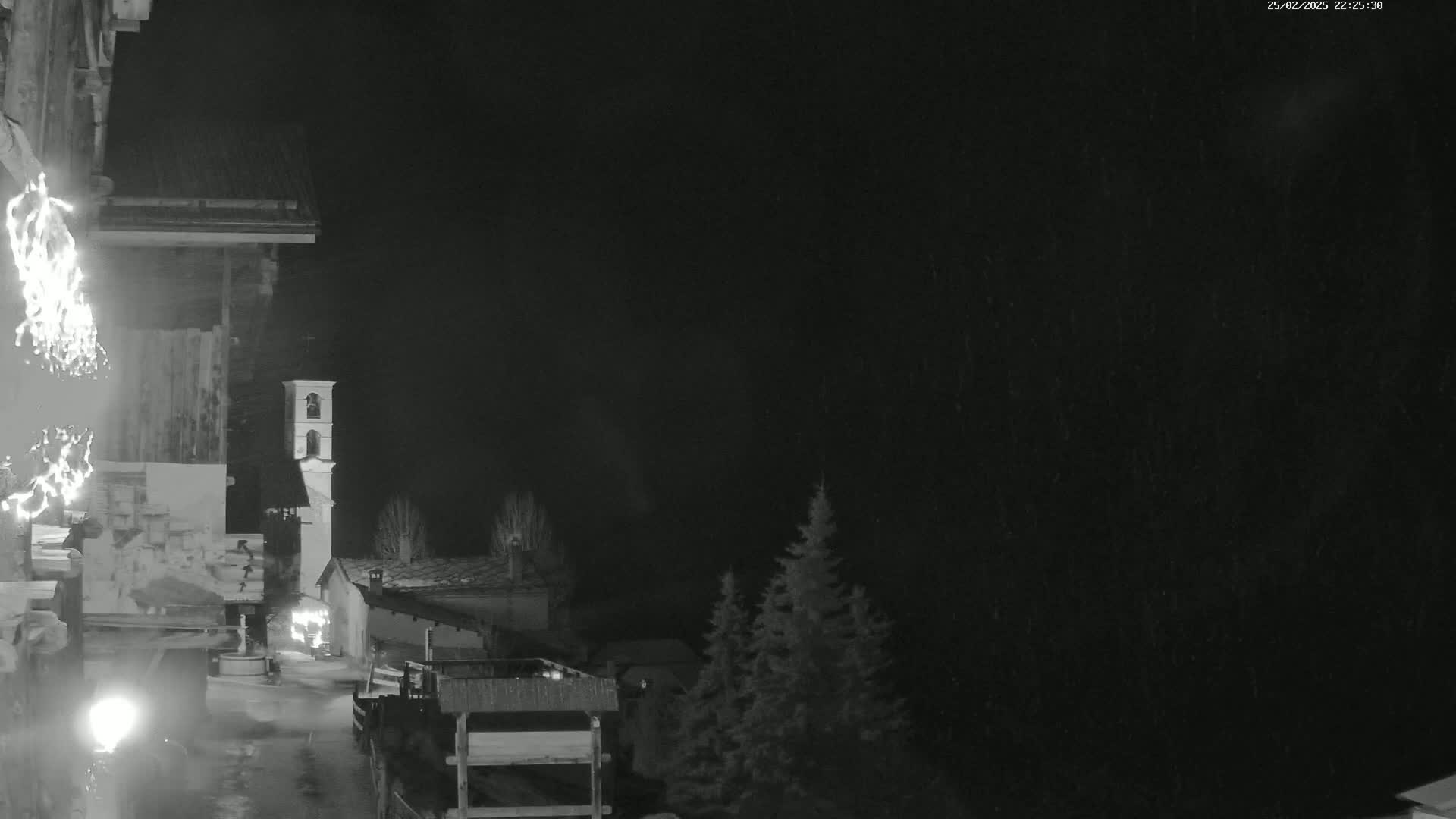 A nighttime, snowy scene shows a village with a bell tower, illuminated by artificial lights, under a dark sky.