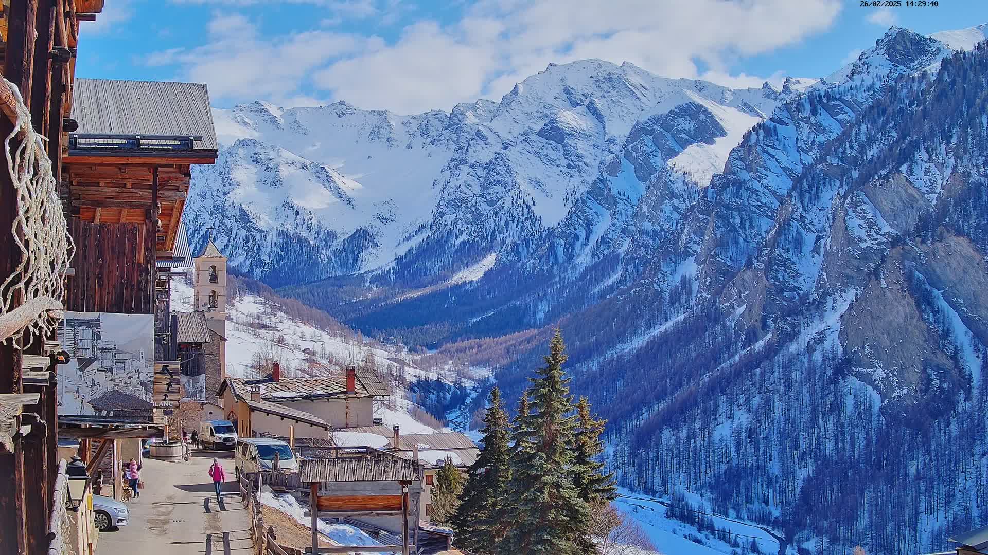 A sunny winter day in a snow-covered mountain village, with a view of towering, snow-capped peaks in the background.