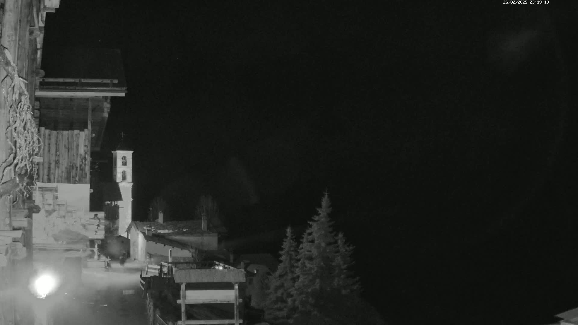 A nighttime, low-light grayscale image shows a village scene with a bell tower, buildings, and evergreens under a dark sky.