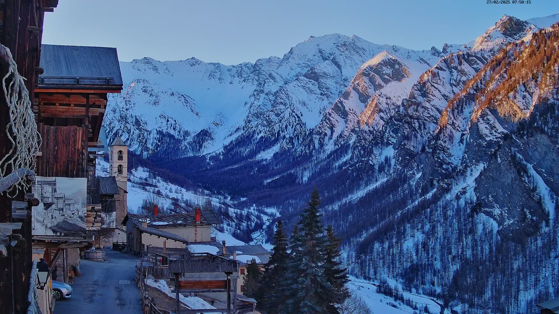 A snow-covered mountain village nestled in a valley, with a clear blue sky and the setting sun illuminating the mountain peaks.