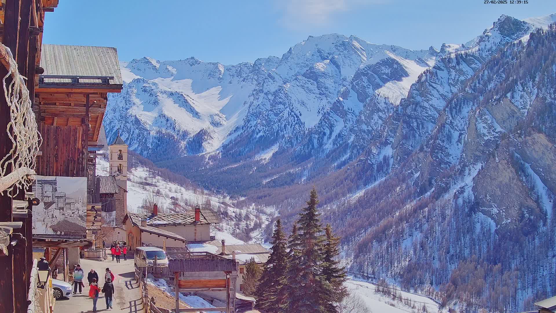 A sunny, snow-covered mountain valley is overlooked by a village with several wooden buildings and people walking on a street.