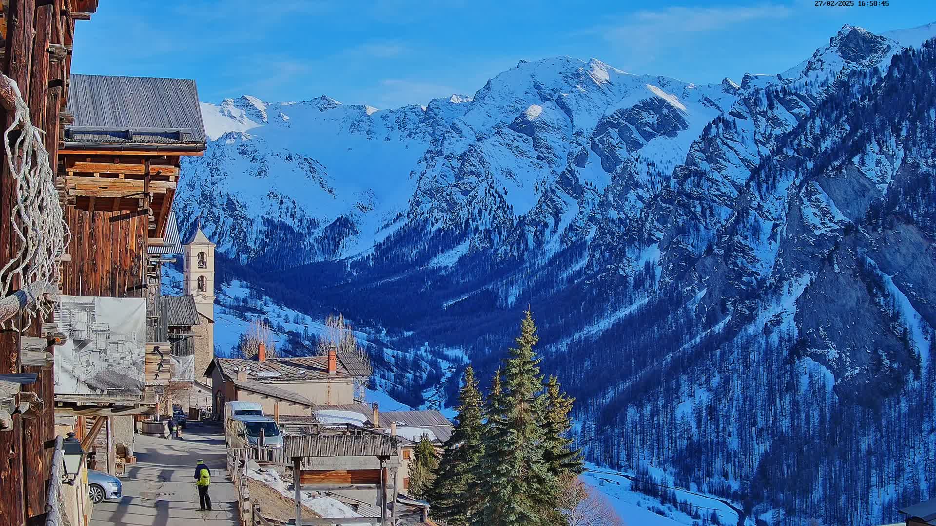 A snow-covered mountain range under a clear blue sky is viewed from a rustic village with wooden buildings, a few cars, and several evergreen trees in the foreground.