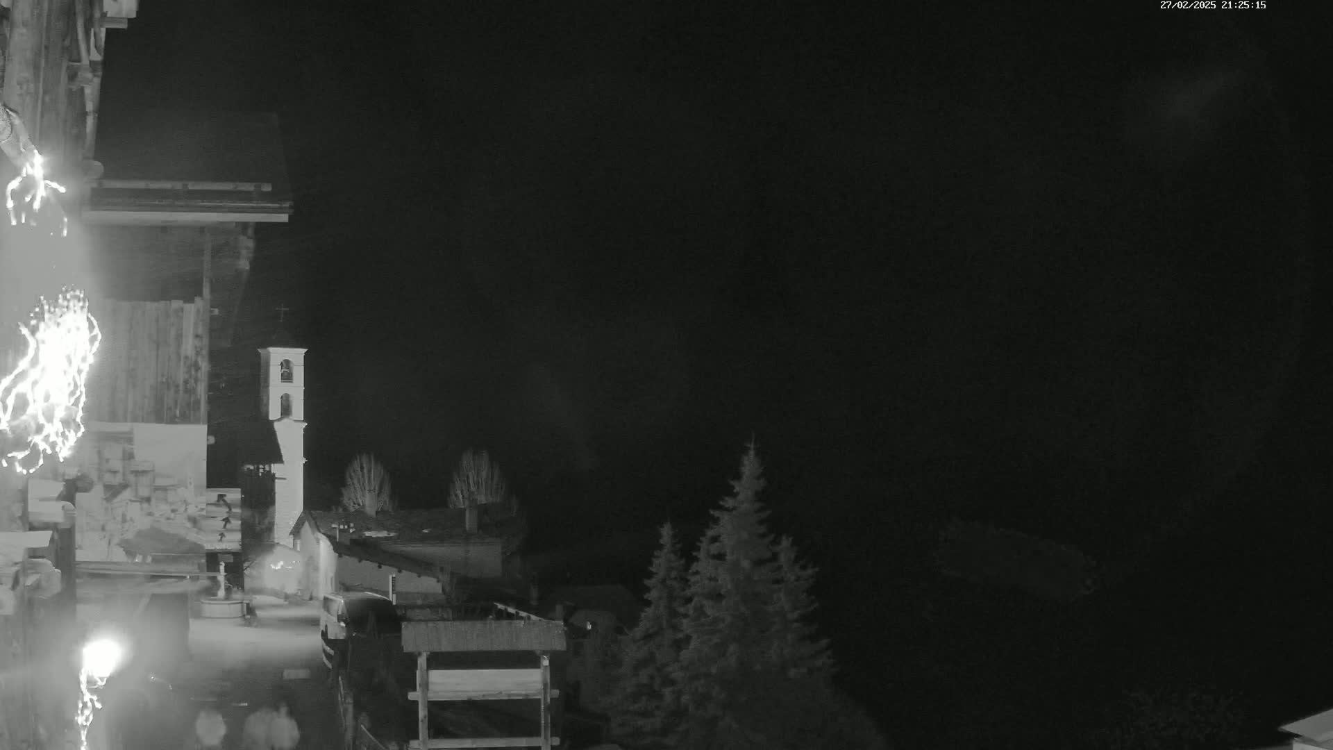 A nighttime, grayscale view of a small village with a church steeple, a few trees, and some buildings, under a dark, possibly cloudy sky.