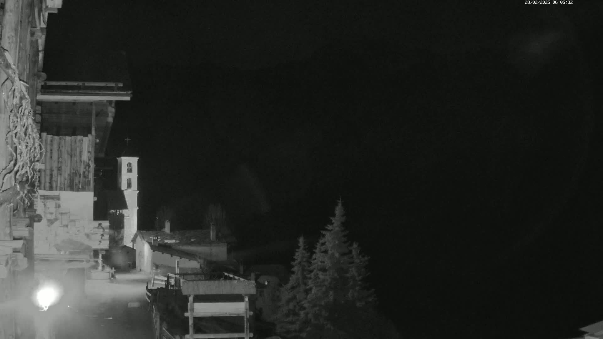 A nighttime, low-light view of a small village with a church steeple, partially obscured by darkness, and a few evergreens in the foreground.
