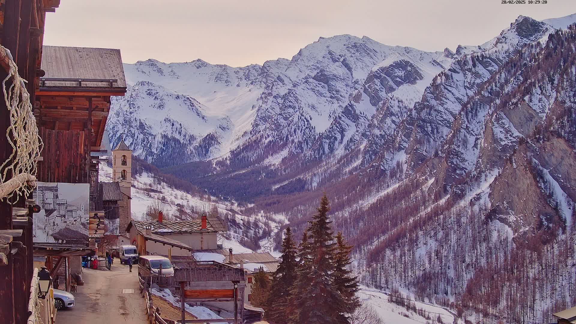 A snow-covered mountain range overlooks a small village nestled in a valley, under a mostly clear sky.