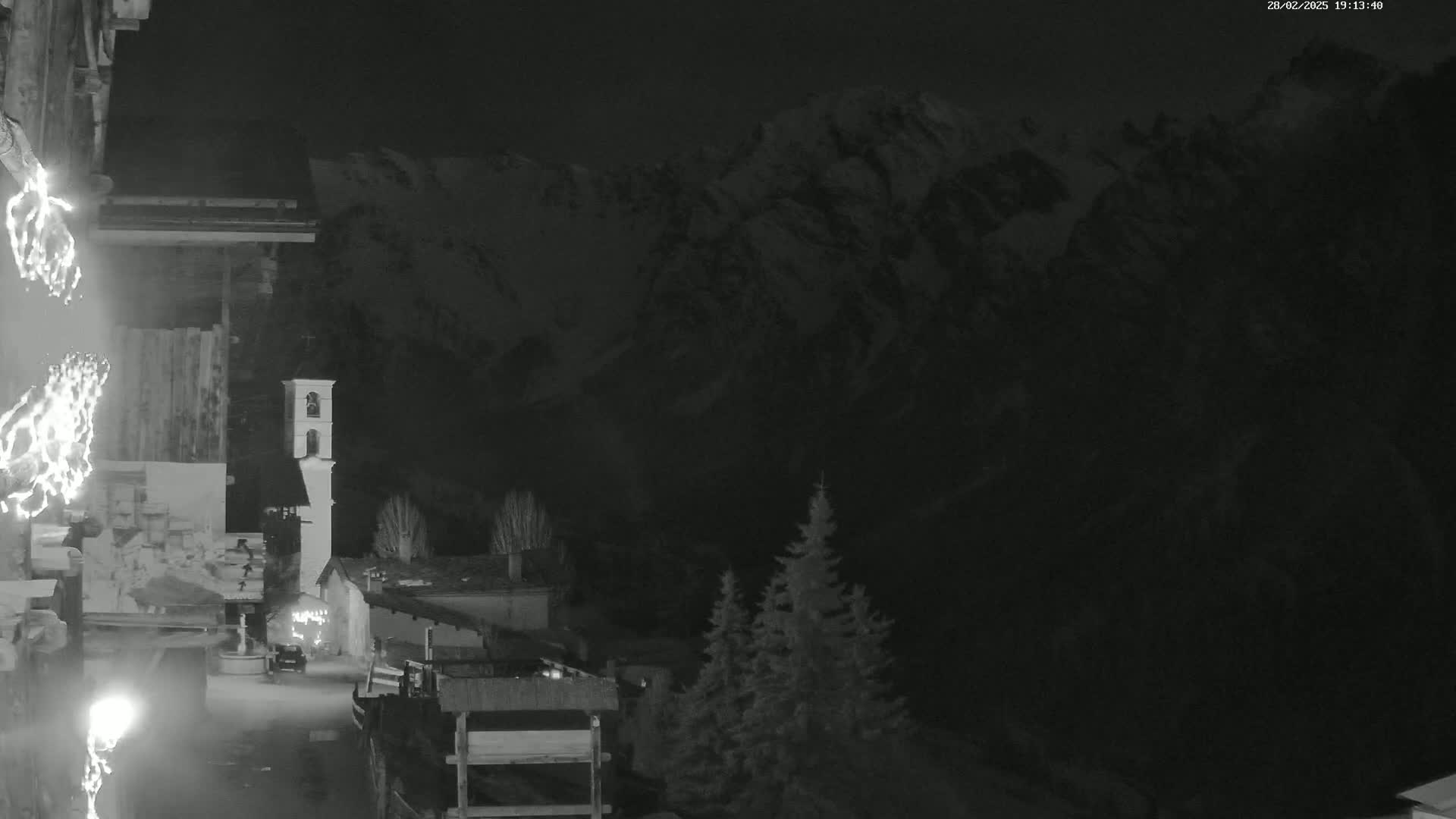 A nighttime, low-light view of a snow-covered mountain range behind a small village with a bell tower, illuminated by artificial lights.