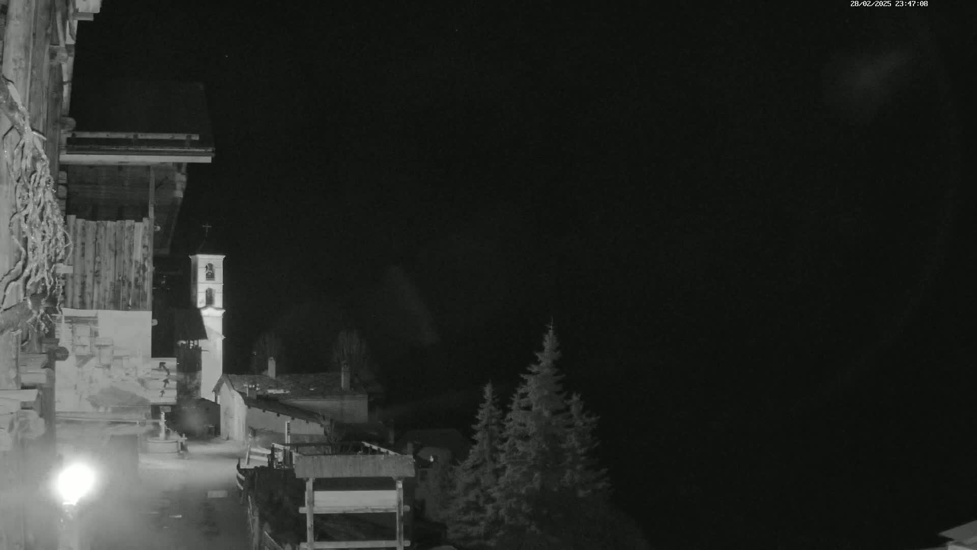 A nighttime, black and white view of a small town or village, featuring a tall bell tower, some buildings, evergreens, and a wooden structure in the foreground under a dark, clear sky.