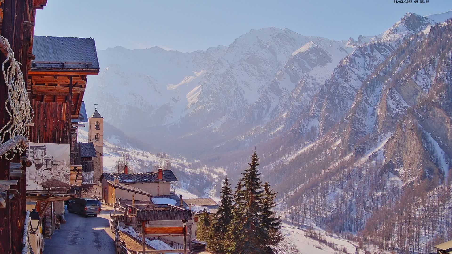A sunny, snow-covered mountain valley is viewed from a village with traditional wooden buildings, a church steeple, and evergreen trees.