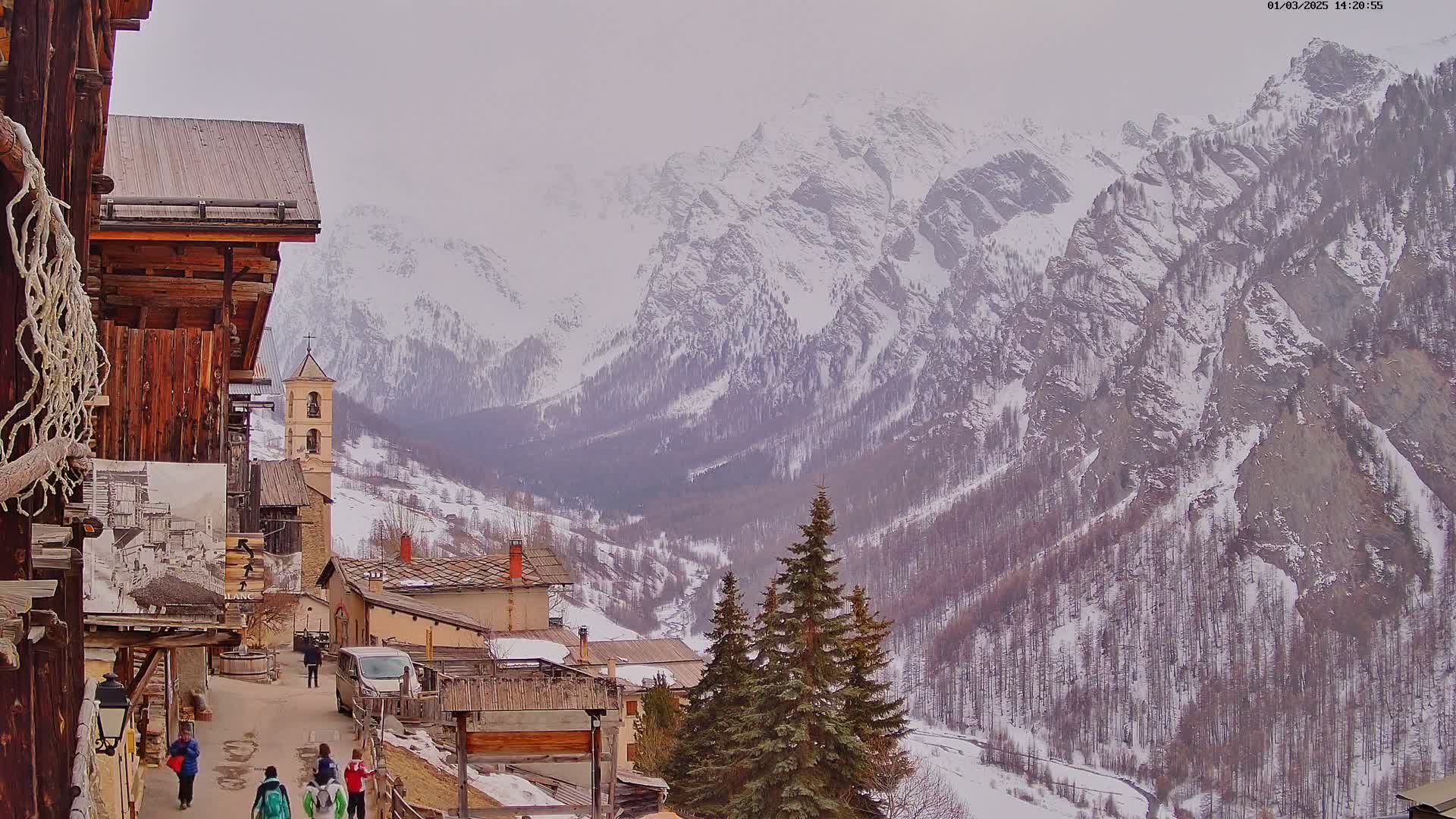 A snow-covered mountain valley is viewed from a village of old wooden buildings, with several people walking along a path.