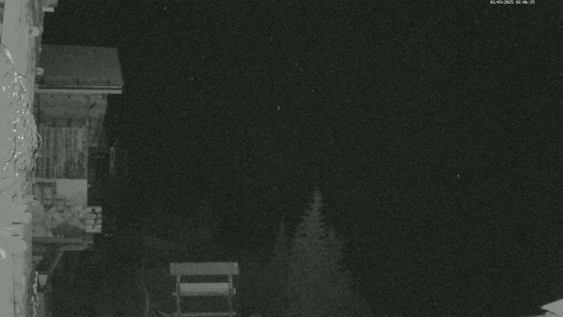 A nighttime view shows a wooden building partially visible, with some trees and a small structure in the background under a dark, possibly snowy sky.