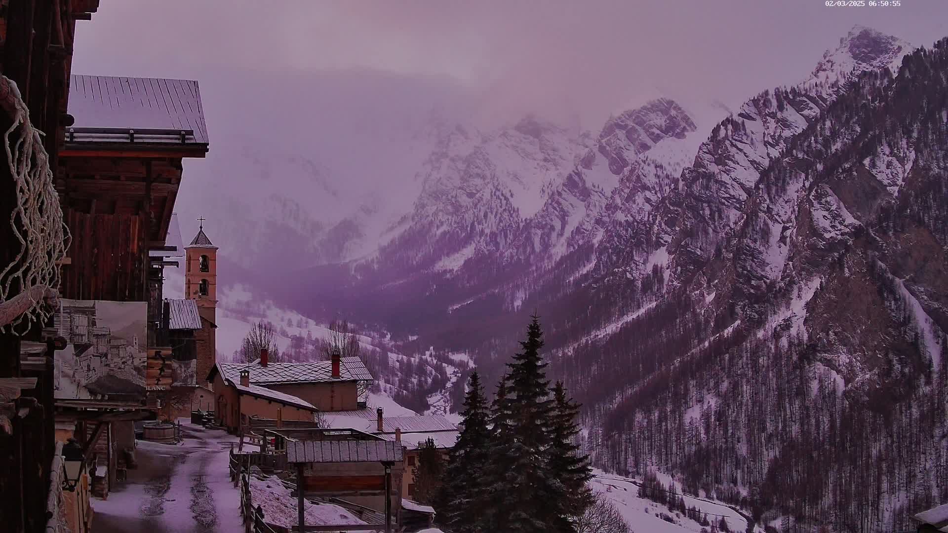 A snow-covered village nestled in a valley, overlooked by towering, snow-capped mountains under a hazy purple sky.