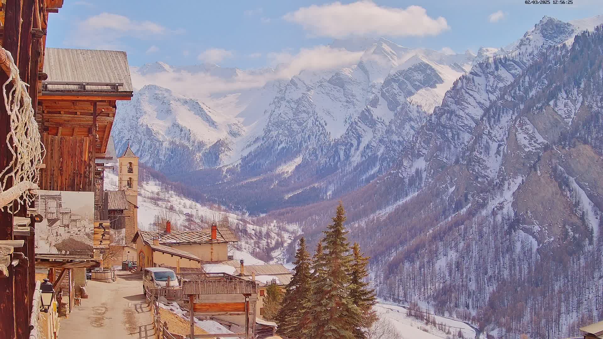 A snow-covered mountain range is visible behind a small village of old stone buildings, under a mostly sunny sky.