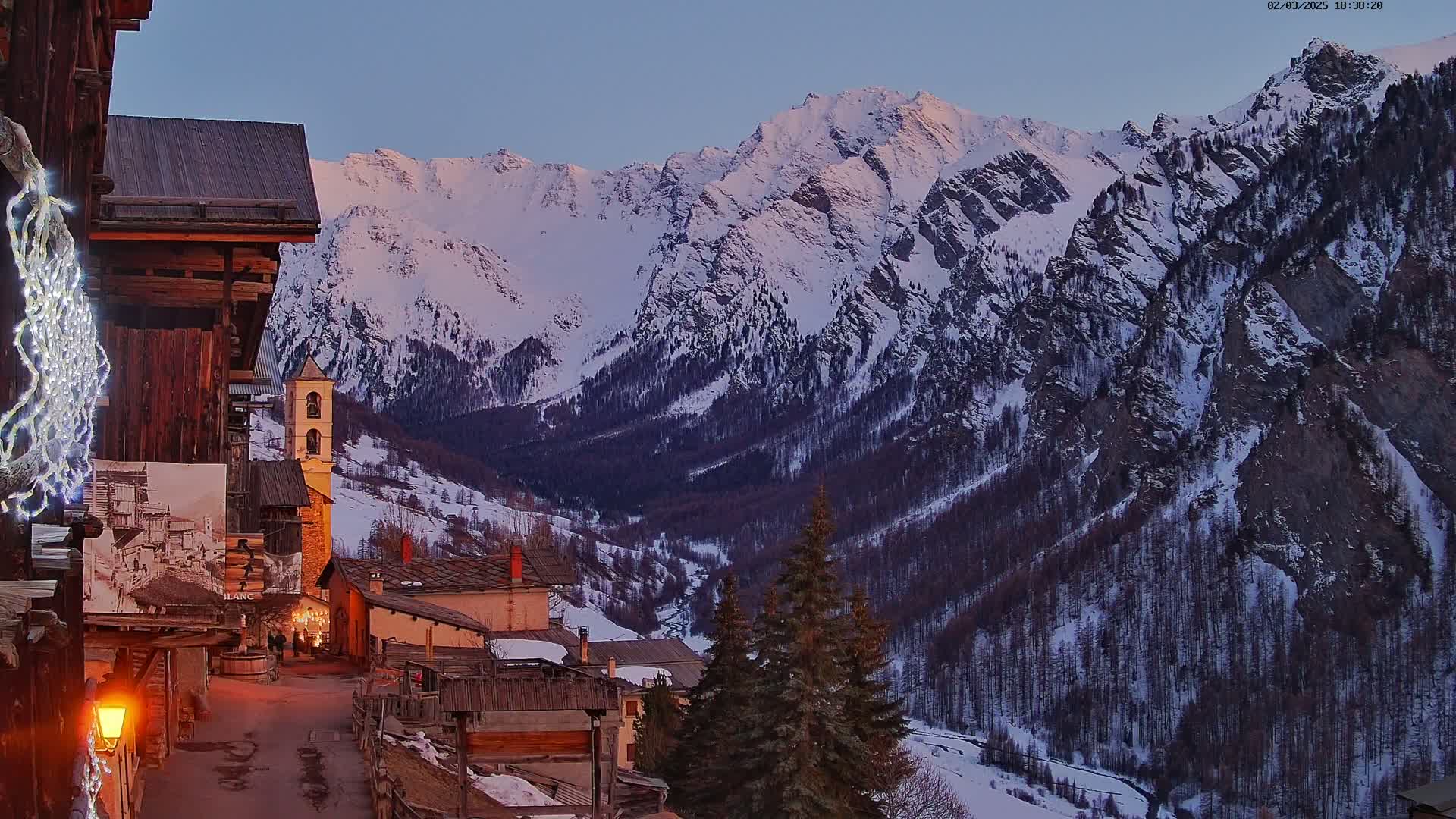 A snow-covered mountain range at twilight overlooks a small village with old wooden buildings and a stone church, illuminated by the soft light of dusk.