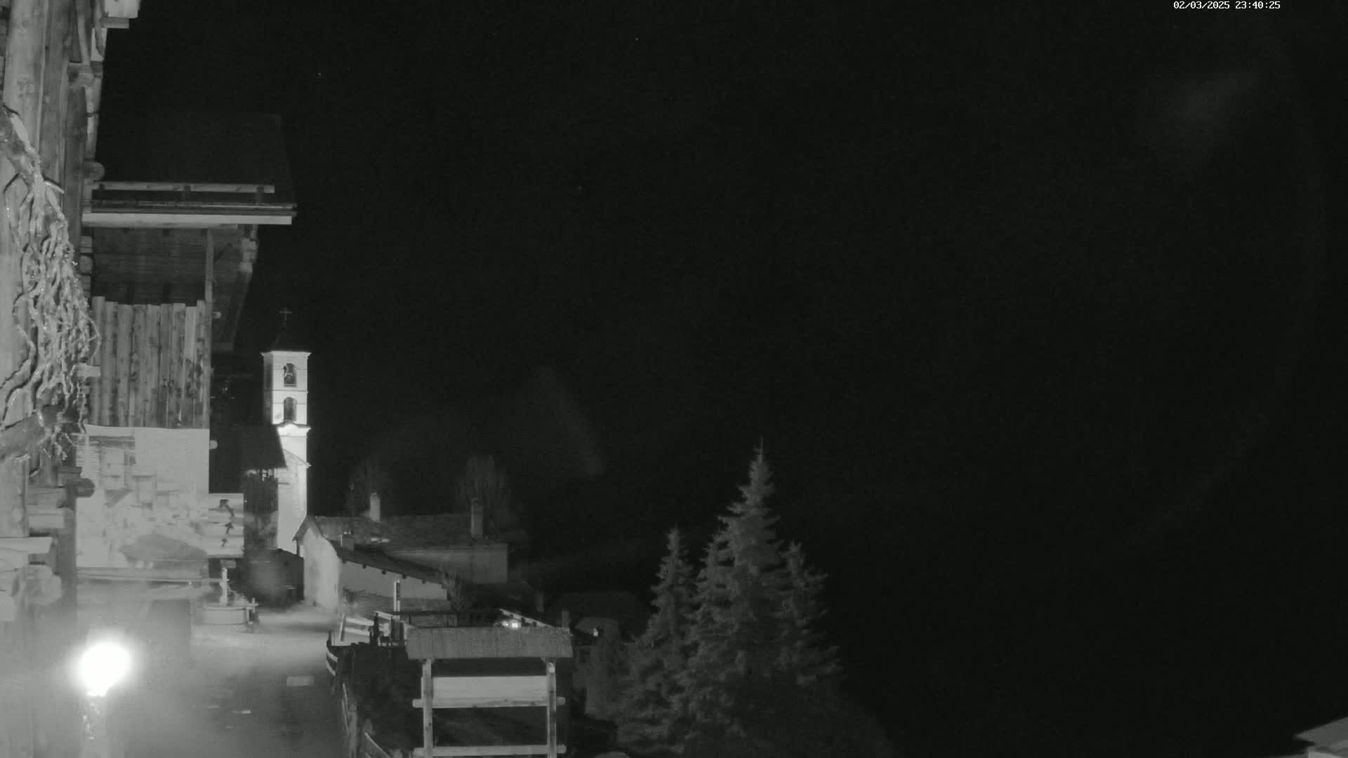 A nighttime, black and white view shows a small village with a church steeple, partially obscured by darkness, and a few evergreens in the foreground.
