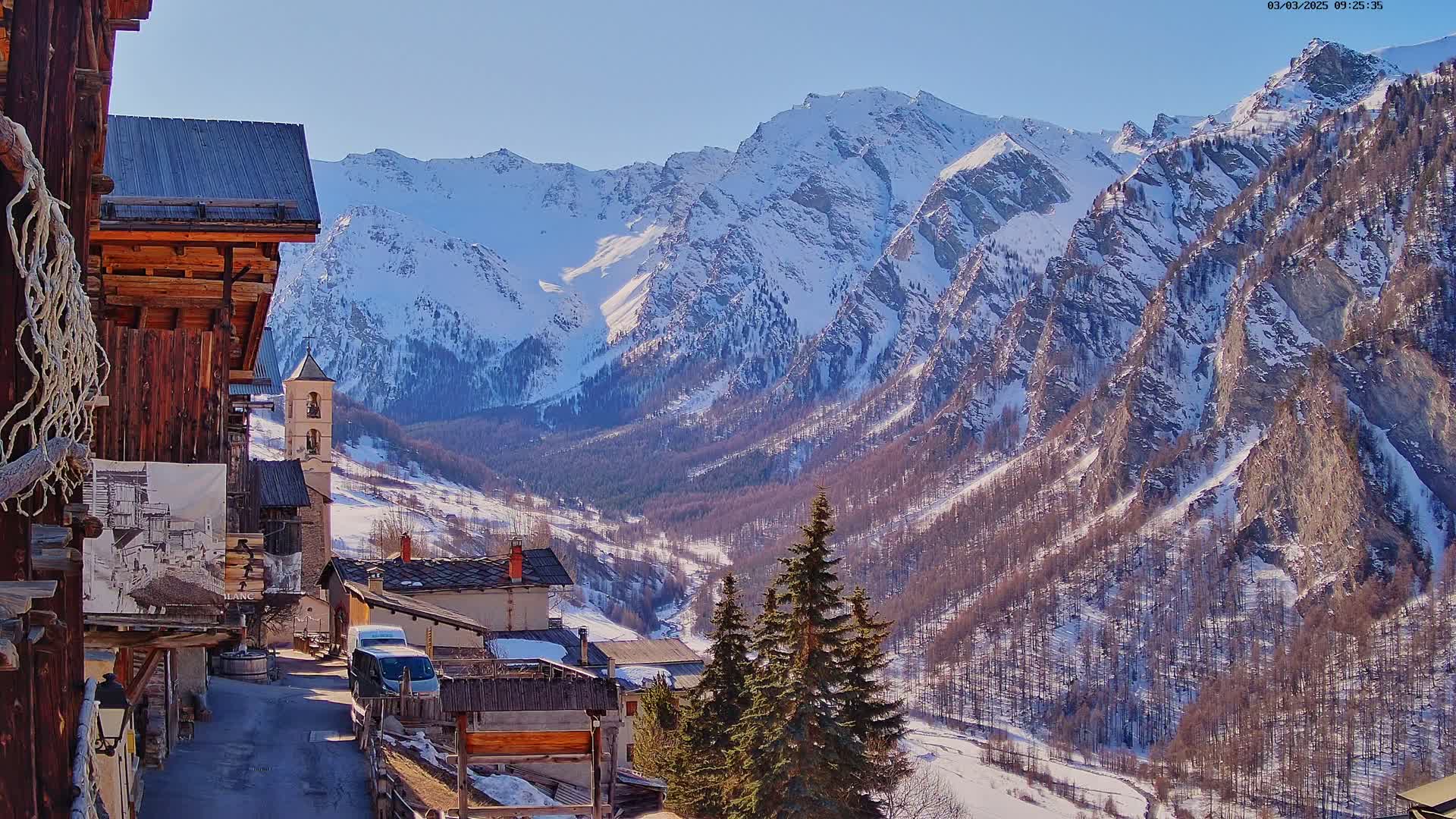 A snow-covered mountain range overlooks a small village of rustic wooden buildings on a sunny day.