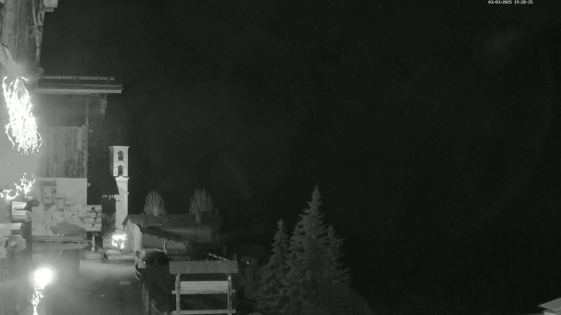 A nighttime, black-and-white view shows a village scene with a bell tower, some buildings, trees, and a vehicle, under a dark, clear sky.