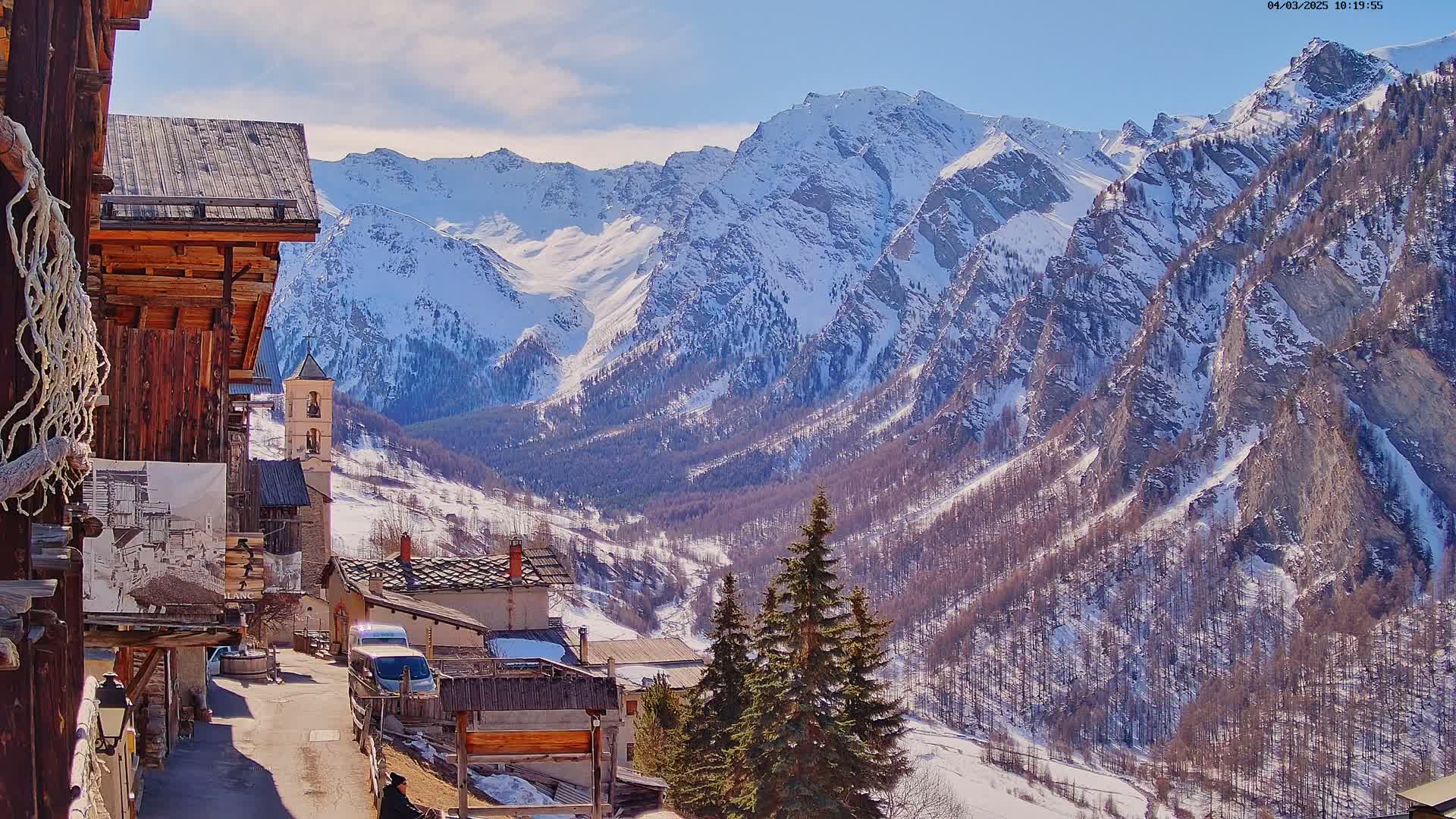 A sunny, snow-covered mountain range overlooks a small village with rustic wooden buildings and a church.