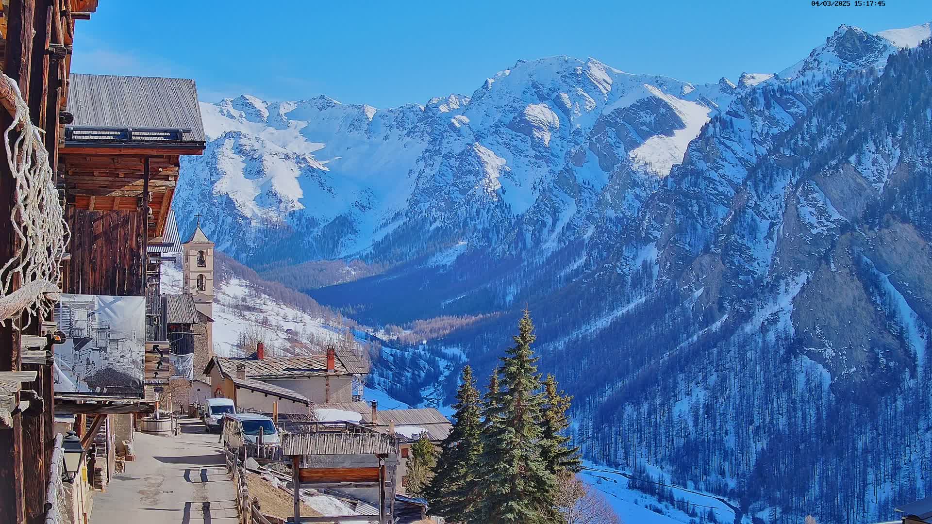A sunny day reveals a snow-covered mountain range overlooking a quaint village nestled in a valley, with several evergreen trees in the foreground.