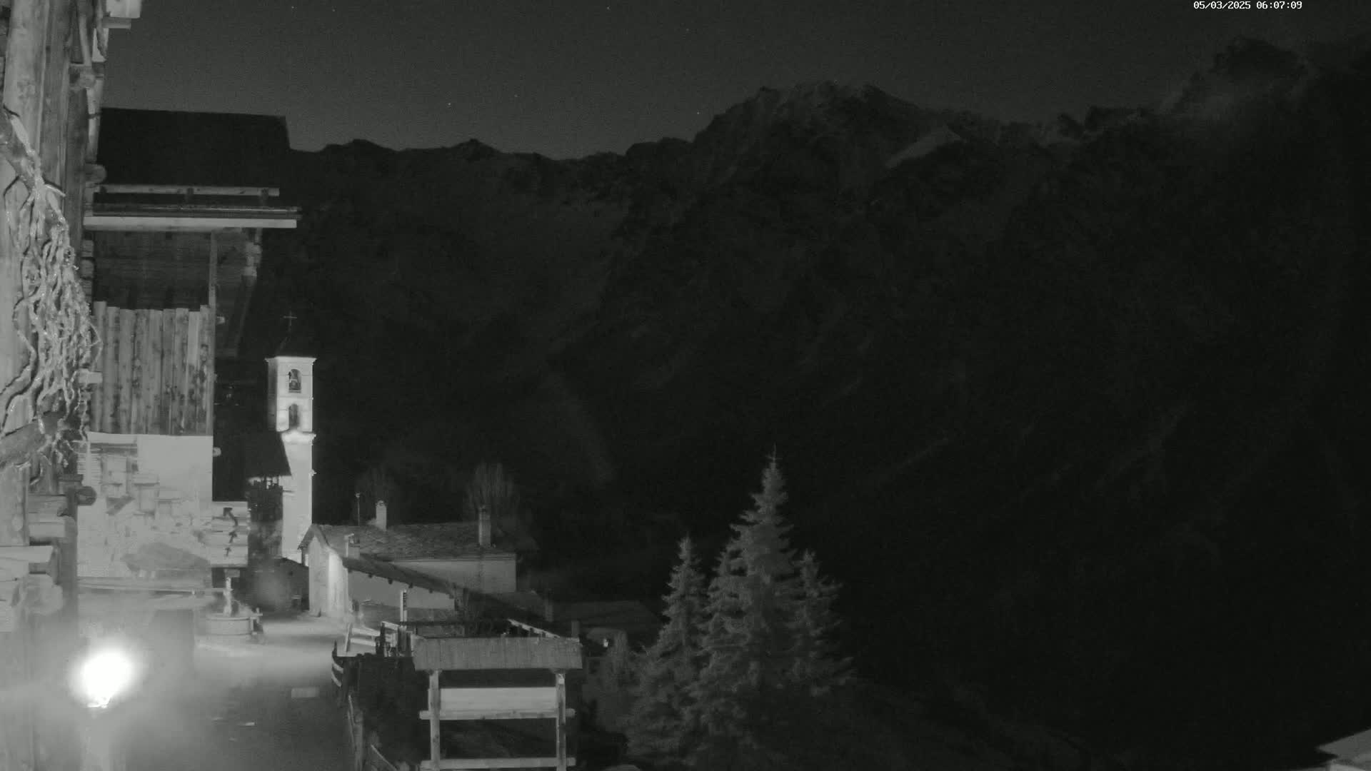 A nighttime, black-and-white view of a village nestled in a valley, with snow-capped mountains in the background under a clear sky.