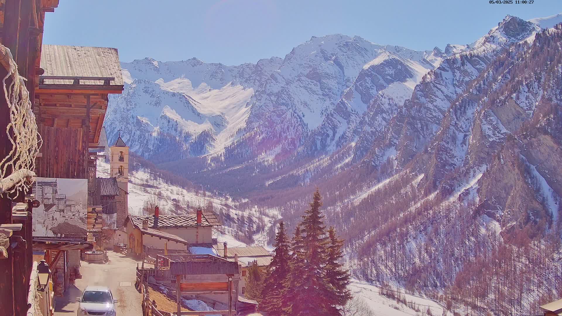 A sunny day reveals a snow-capped mountain range overlooking a small village nestled in a valley, with evergreen trees dotting the landscape.