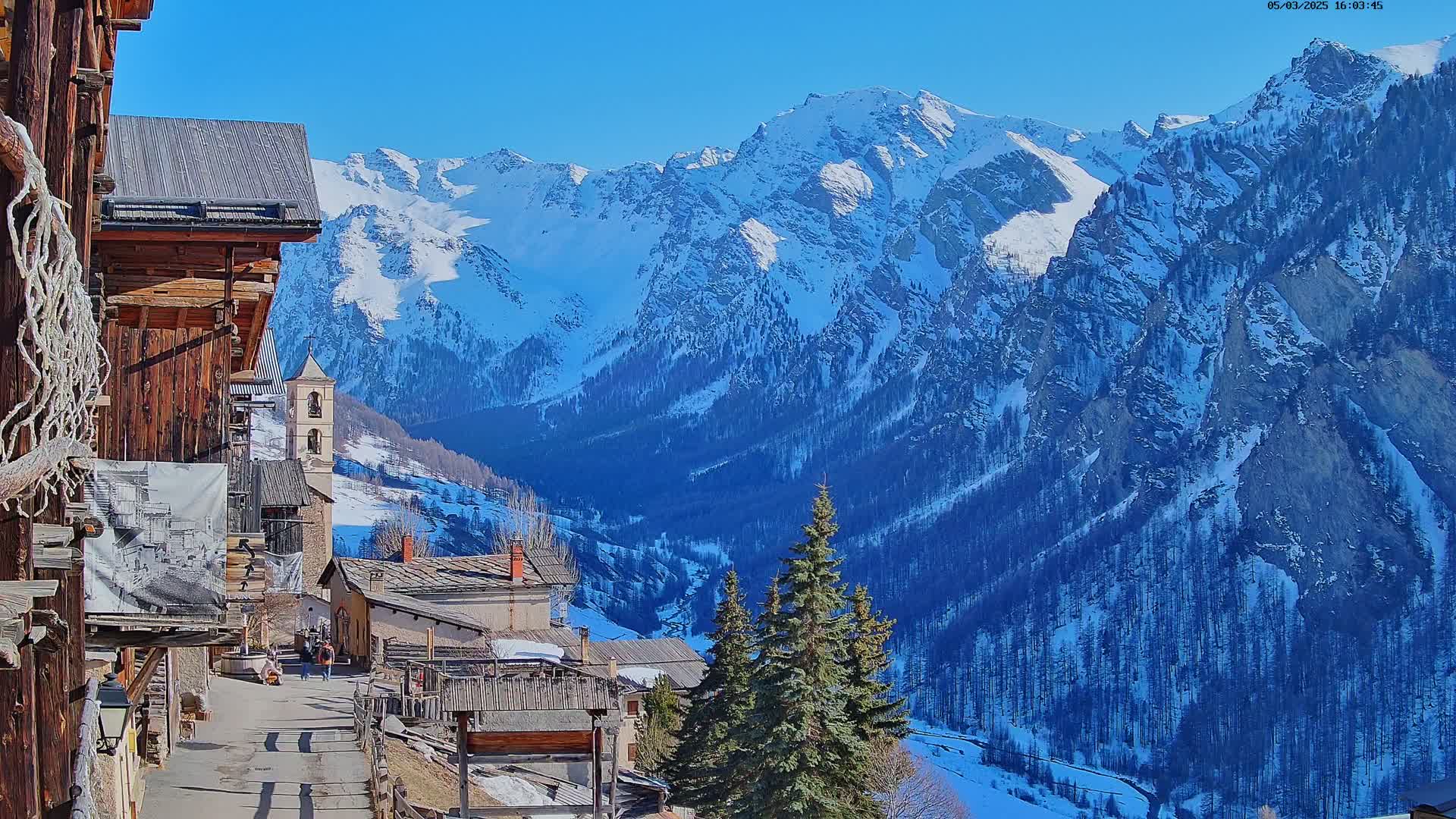 A quaint village nestled in a valley, with snow-covered mountains rising in the background under a clear blue sky.