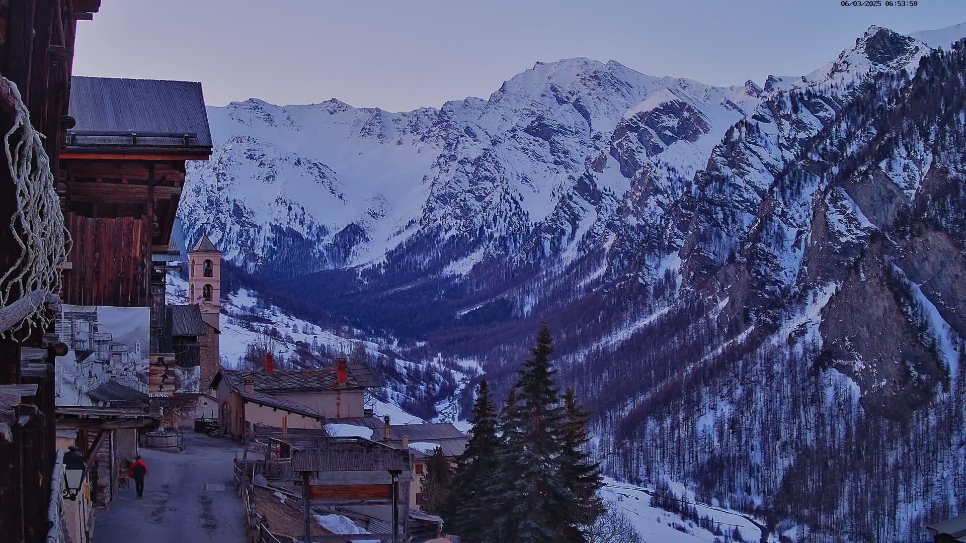 A snow-covered mountain range is viewed from a village street at dusk, with a few evergreen trees in the foreground.