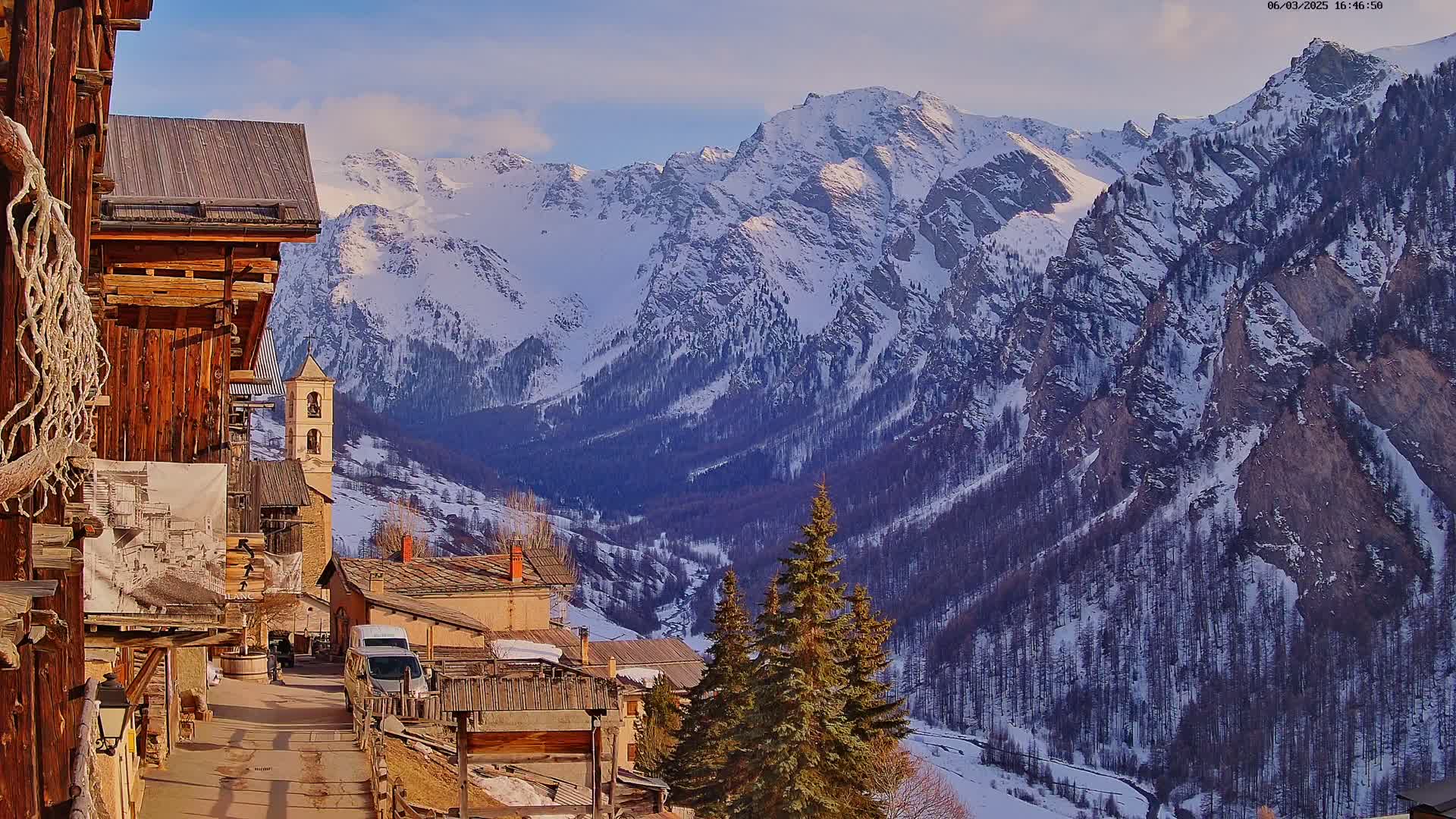 A snow-covered mountain range is seen in the background from a village with rustic wooden buildings, under a mostly clear sky.