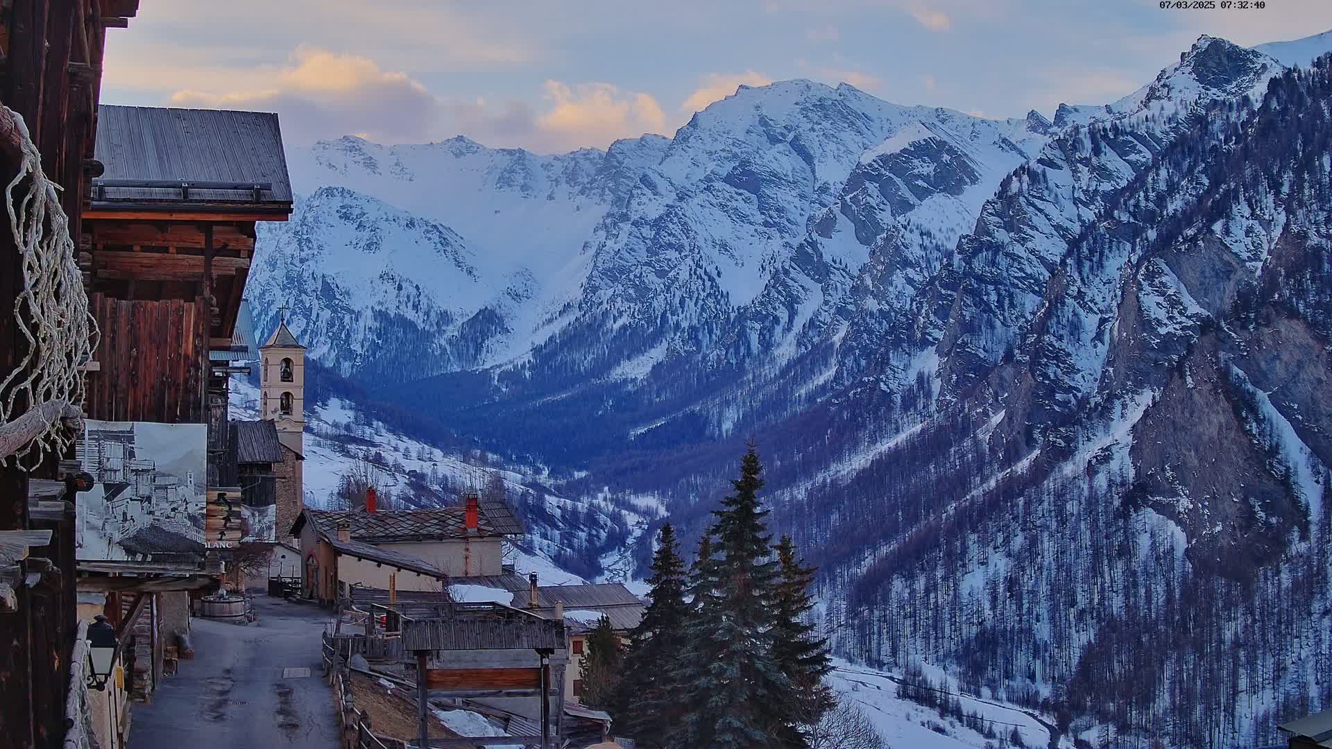A snow-covered mountain range is visible behind a small village of rustic buildings, under a partly cloudy sky.