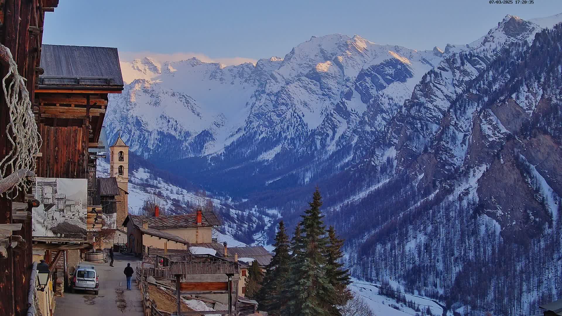 A snow-covered mountain range is viewed from a village of rustic wooden buildings under a clear, twilight sky.
