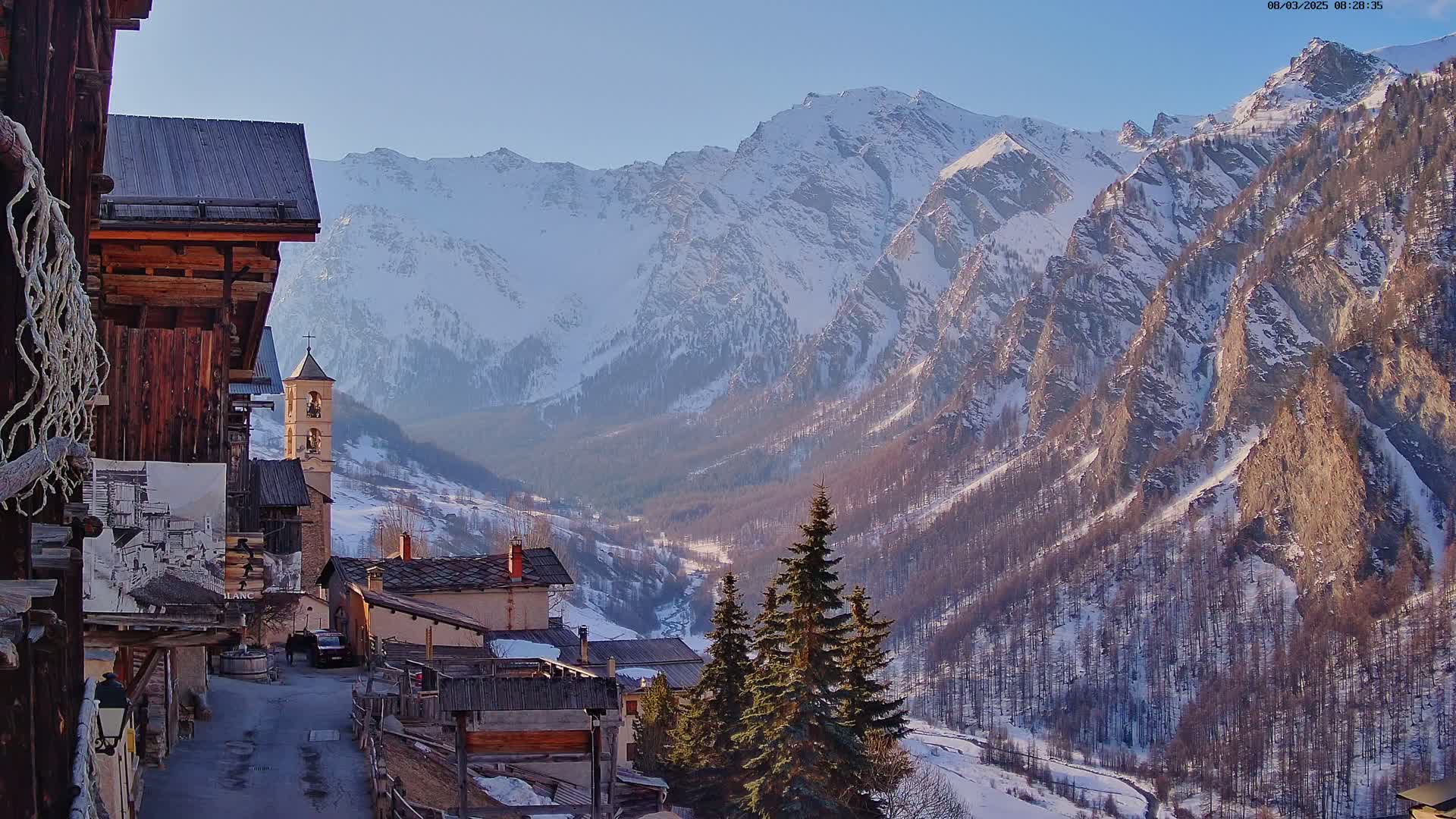 A quaint mountain village nestled in a valley, overlooked by snow-capped mountains under a clear blue sky.