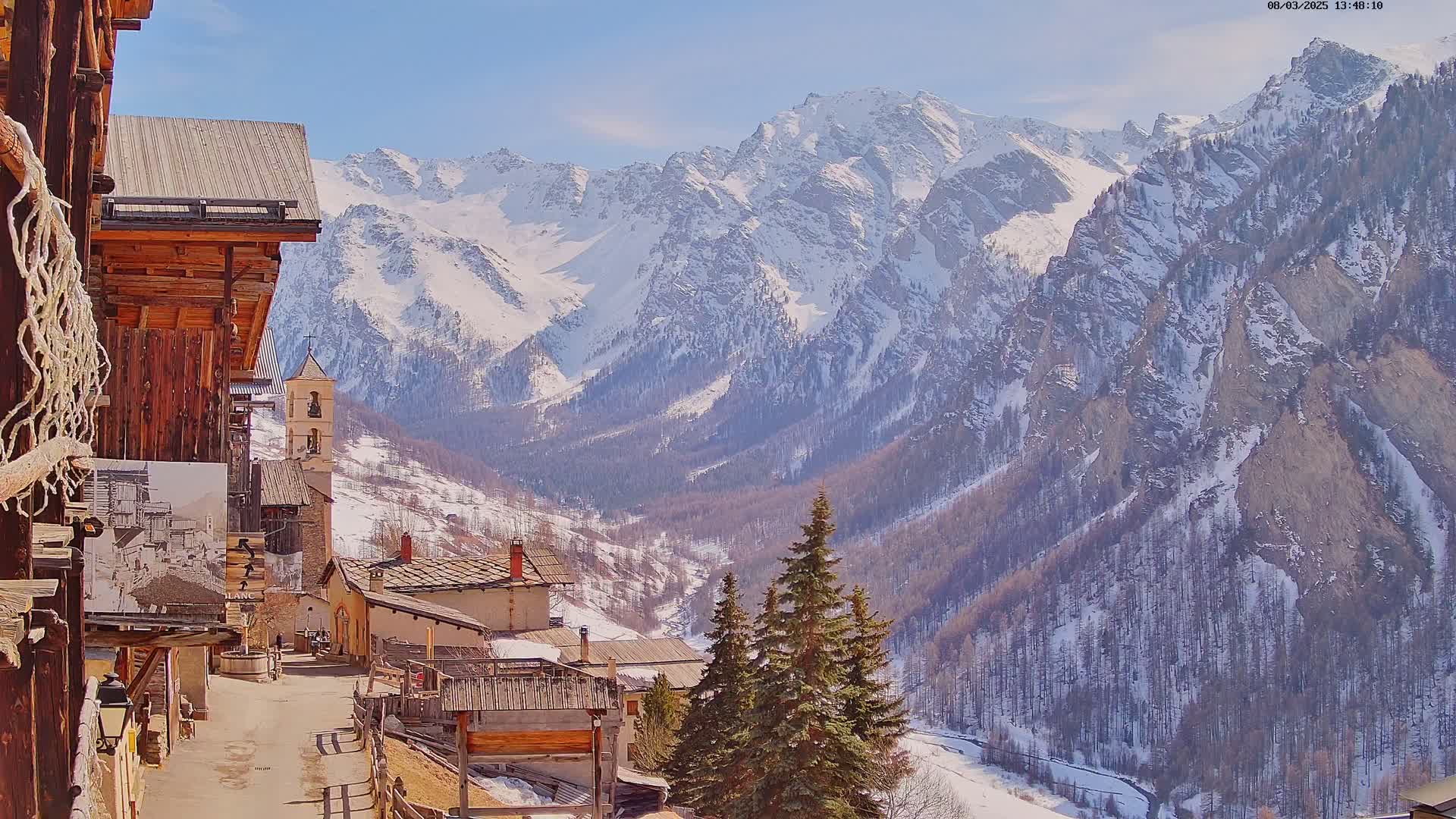 A sunny view of a snow-covered mountain valley with a small village nestled amongst wooden buildings, a church steeple, and evergreens in the foreground.