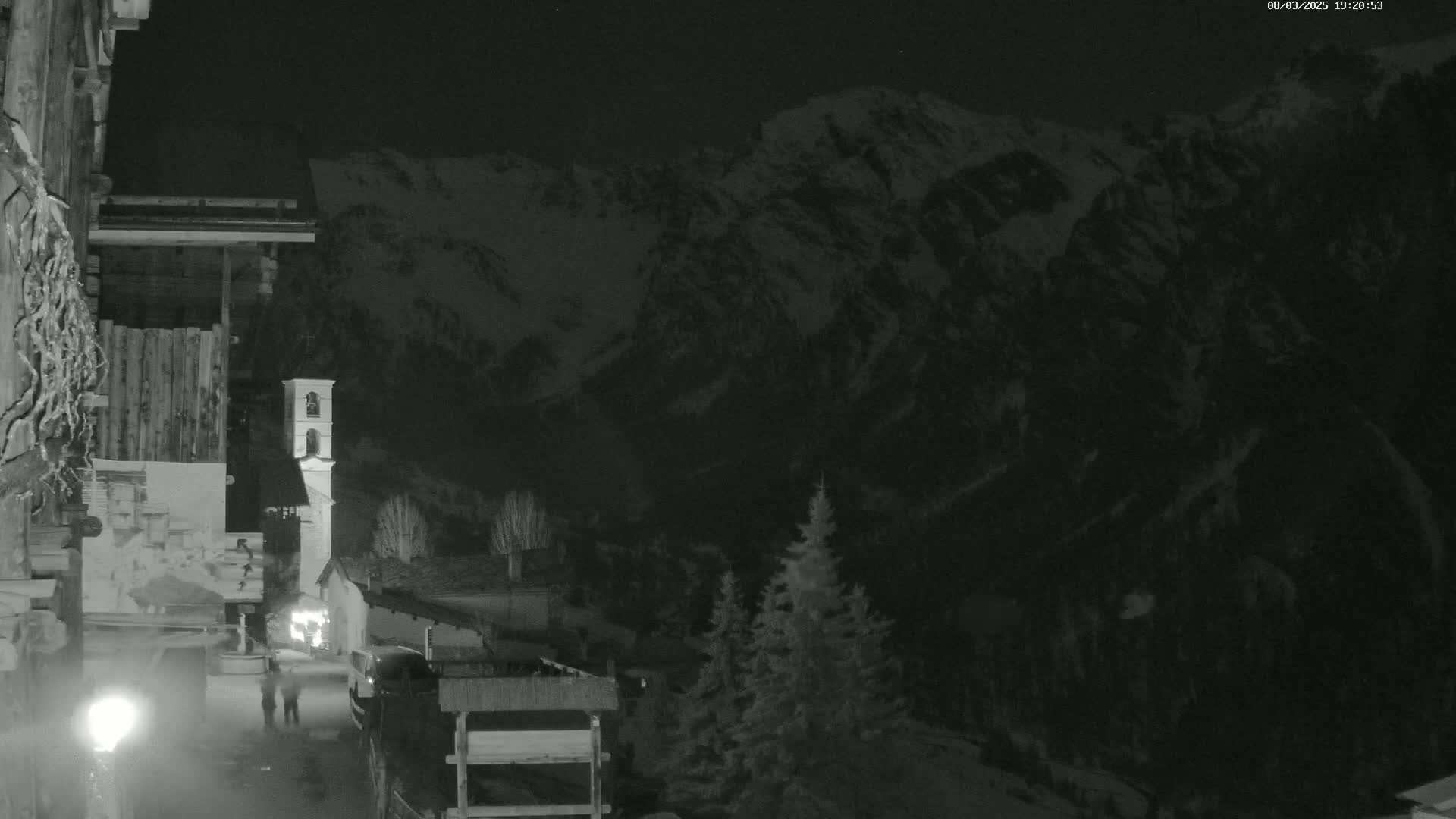 A nighttime view of a village nestled in a valley, overlooked by snow-capped mountains under a dark sky.