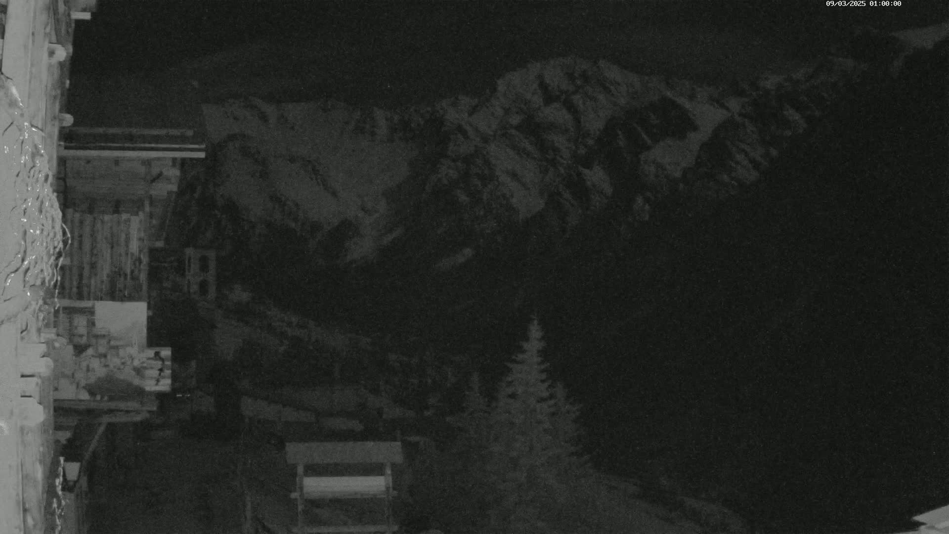 A nighttime, low-light view shows snow-covered mountains in the distance, with a dark, wooded area in the foreground, partially obscuring a building.