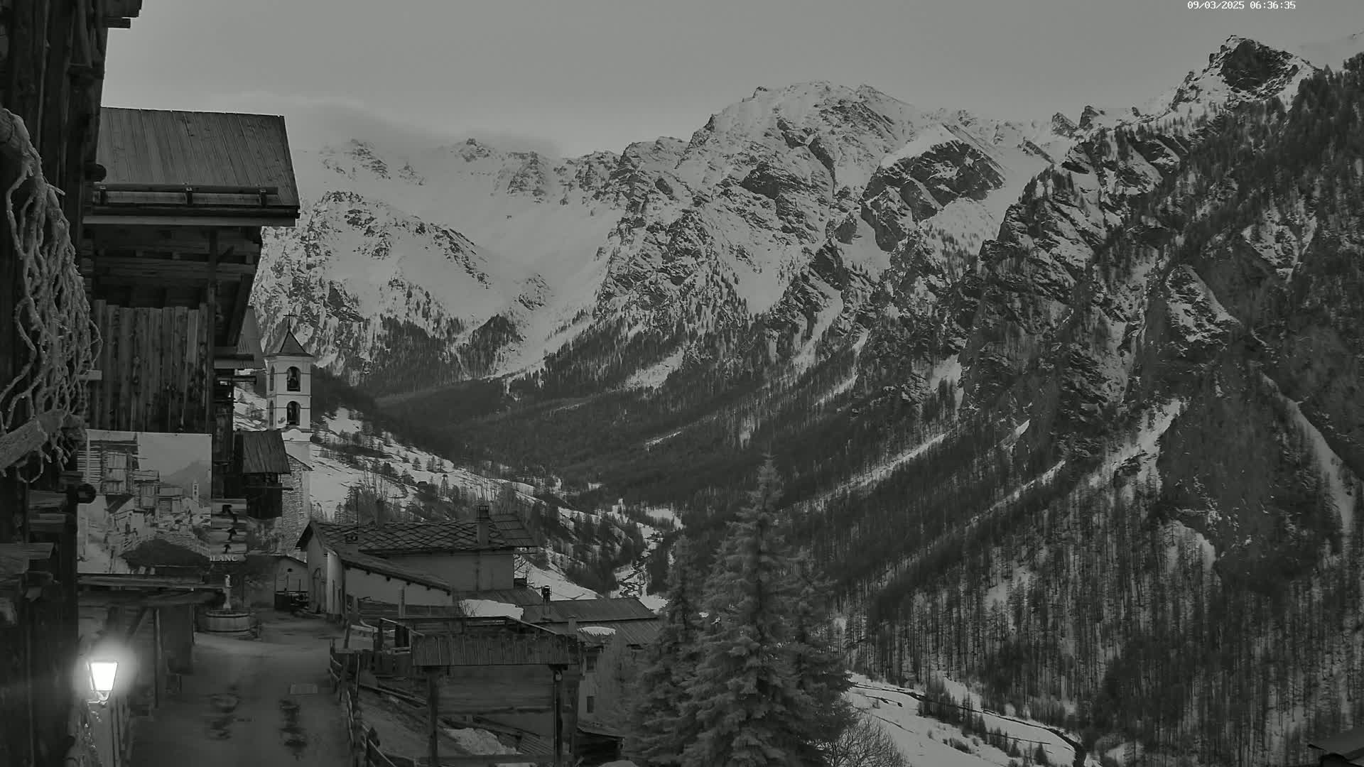 A snow-covered mountain range is viewed from a village street at twilight, with a church steeple visible in the mid-ground.