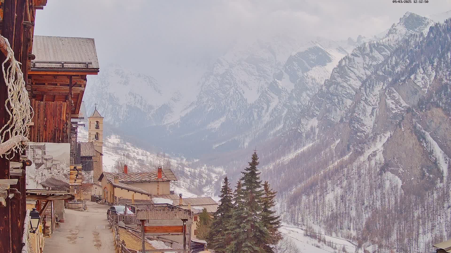 A snow-covered mountain valley is seen from a village of rustic stone and wood buildings under an overcast sky.