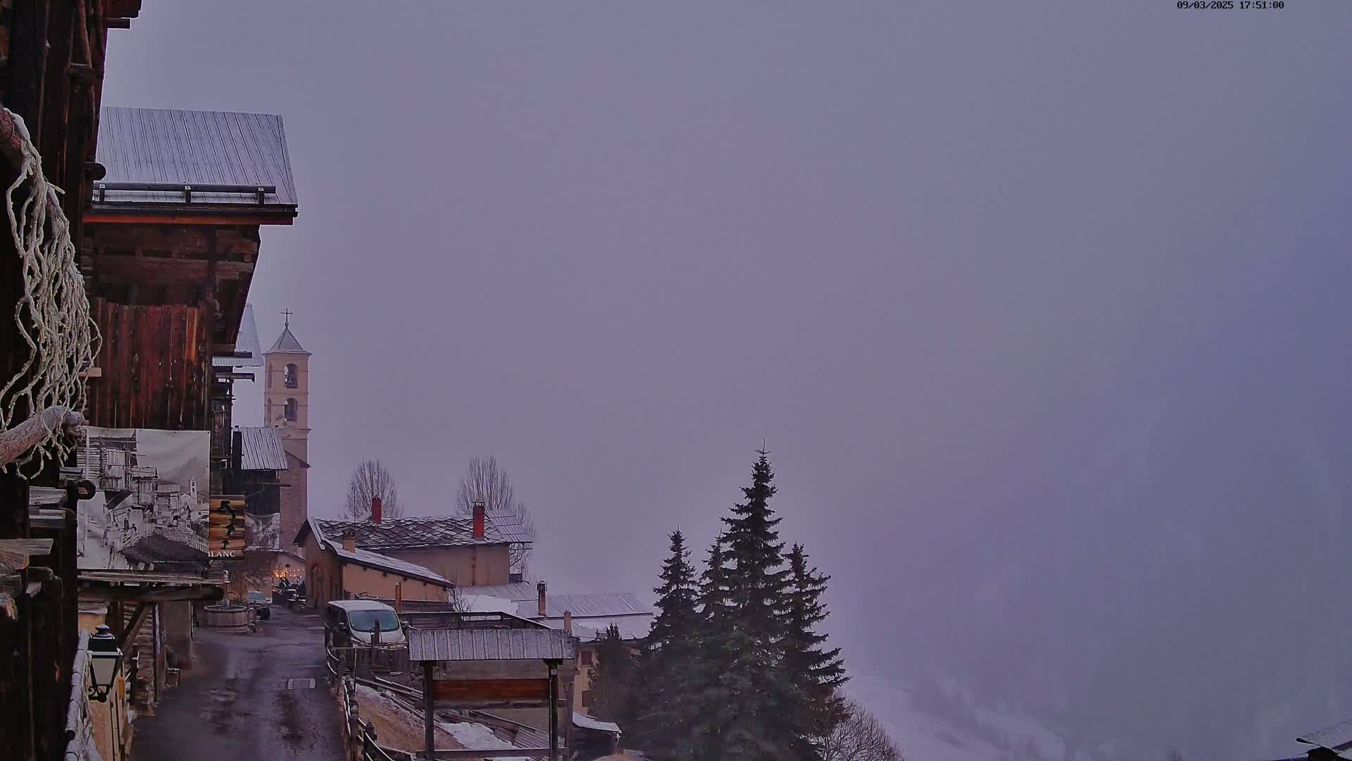 A foggy, overcast view of a village nestled in a mountainside, featuring traditional buildings and a church steeple.